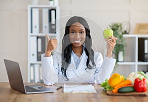 Millennial black dietitian holding apple and showing thumb up gesture at desk in medical office. Healthy eating concept