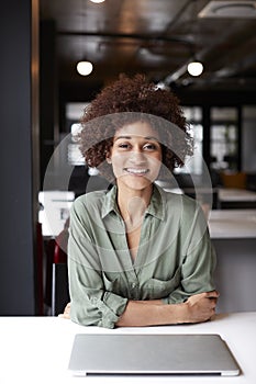 Millennial black female creative sitting at desk in an open plan office smiling to camera, vertical
