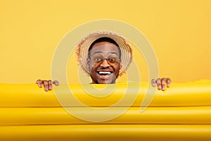Millennial black guy in straw hat and sunglasses hiding behind inflatable lilo on yellow studio background