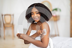 Millennial black lady applying cream, lotion or body milk after bath, making domestic beauty procedure indoors