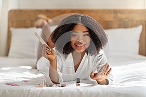 Millennial black lady doing manicure, holding nail file, smiling at camera, lying on bed at home. Beauty and wellness