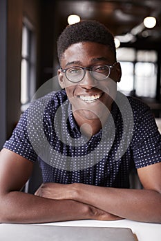 Millennial black male creative sitting at a desk in an office smiling to camera, close up, vertical