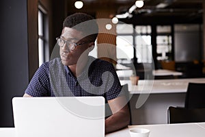 Millennial black male creative sitting in an office using laptop, looking out of window, close up