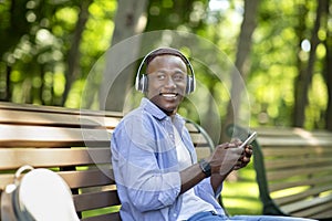 Millennial black man sitting on bench in park and listening to music from mobile phone