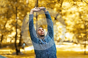 Millennial black sportsman stretching his arms before jogging at park on autumn morning