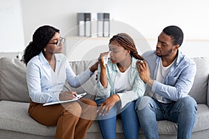 Millennial black woman crying at psychologist's office, loving husband supporting her with professional counsellor