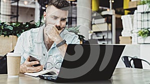 Millennial businessman sitting in cafe with open laptop, looking thoughtfully at computer screen, holding smartphone. photo