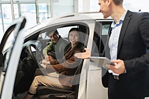 Millennial car salesman discussing purchase of new auto with happy young couple after test drive at dealership centre