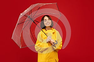 Millennial caucasian woman posing with transparent umbrella, smiling and looking at camera over red background