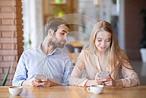 Millennial couple having dull date, stuck in smartphones, jealous young man looking into woman`s gadget at cafe