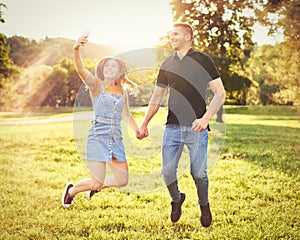 Millennial couple holding each other`s hand and jumping together outdoors in a park while taking selfie - Happy people lifestyle