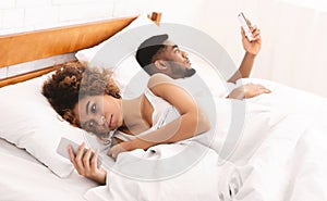 Millennial couple lying on bed back to back with smartphones
