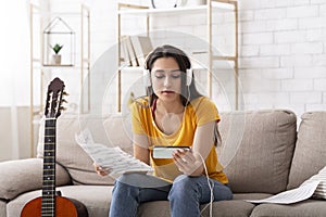 Millennial girl in headphones learning to play guitar by watching online video tutorial on mobile phone, indoors