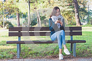 Millennial girl is sitting on a bench in a park and looking her smartphone with smiling and surprised expressio