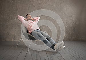 Millennial Guy Relaxing Sitting In Comfortable Chair Holding Hands Behind Head Resting With Eyes Closed Over Gray Wall