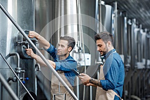 Millennial guy worker in apron turns a valve on big boiler for lager, manager with tablet controls process in interior