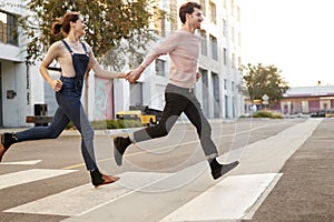 Millennial hipster couple running across the road in the city holding hands, side view