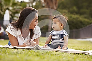 Millennial Hispanic mother lying beside her baby, sitting on a blanket in the park, close up