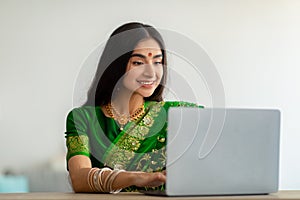 Millennial Indian lady in stylish sari dress studying or working online, using laptop at home
