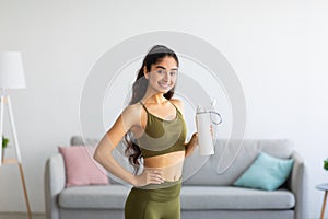 Millennial Indian woman in sportswear drinking water or protein shake from bottle at home