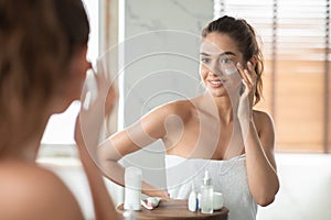 Millennial Lady Applying Facial Moisturizer On Cheek Posing In Bathroom