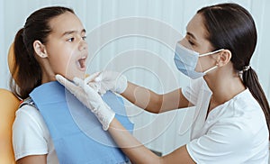 Millennial lady doctor in uniform, protective mask and rubber gloves examines of small child with open mouth