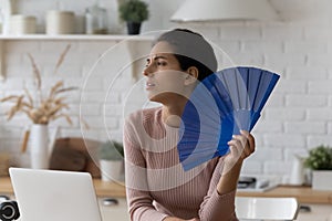 Millennial latin female freelancer use hand fan in unventilated kitchen