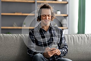 Millennial Man Enjoying Music Using Phone Wearing Headphones At Home