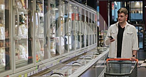 Millennial man pushing shopping cart and looking at phone screen near supermarket fridges. Young guy using smartphone