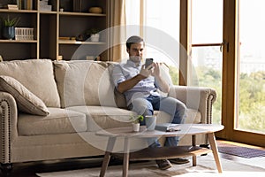 Millennial man wearing wireless headphones relaxing on sofa using phone
