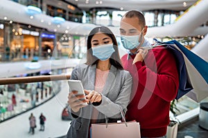 Millennial multiracial couple in face masks checking shopping list in mobile app, holding shopper bags at mall