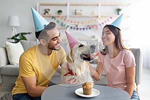 Millennial multiracial couple in party hats celebrating pet dog& x27;s birthday with small festive cake at home