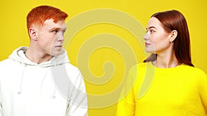 Millennial redhead man and woman looking at camera and turning to each other in slowmo at yellow background. Portrait of