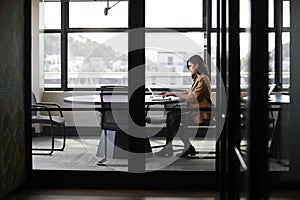 Millennial white creative businesswoman working alone in a meeting room, seen through glass wall