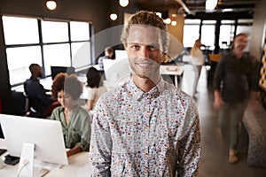 Millennial white male creative standing in a busy casual office, smiling to camera