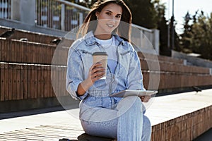 Millennial woman in denim clothes sits on park bench, holding a smart tablet in her hands. Woman uses tablet