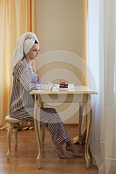 millennial woman in towel on her hand and pijamas on her body sit at table read interesting book. drink tea with cake in