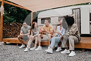 Miltiethnic young friends sitting near motorhome outdoors, talking and laughing, enjoying conversation on camping trip