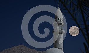 Minaret of a mosque in Jordan with dark blue evening sky and a full moon in the background. Elements of this image furnished by