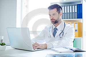 Minded successful doctor sitting at his office and using laptop