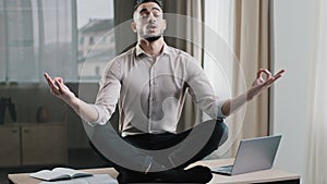 Mindful calm arabian business man employee in lotus position sit on work desk closed eyes meditating take break at home