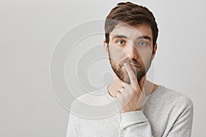 Mindful and calm good-looking male with beard holding index finger over mouth and standing over gray background