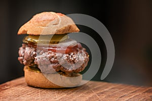 Mini burger with bun, mustard, beef patty, ketchup, onion and gherkin on a rustic wooden kitchen board against a dark background,