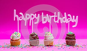 Mini cakes with icing and colorful sprinkles on pink background, text happy birthday decoration