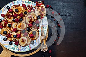 Mini pancakes with berries on a patterned plate with silver spoon in rustic style. Traditional breakfast on black table