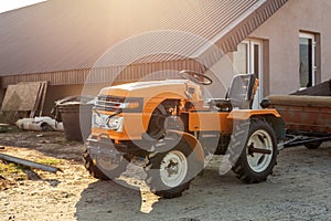 Mini red tractor with trailer standing near hangar building at farm during sunset or sunrise. Small agricultural