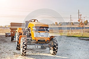 Mini red tractor with trailer standing near hangar building at farm during sunset or sunrise. Small agricultural