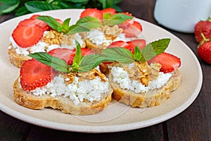 Mini sandwiches with cottage cheese, fresh strawberries, decorated with mint leaves on rye bread on a dark wooden background.