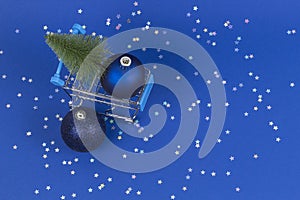 Mini shopping cart with small Christmas tree and navy Christmas decoration bauble balls on blue background with silver