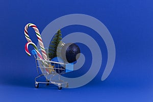 Mini shopping cart with small Christmas tree, Xmas decoration bauble balls and candy canes on navy blue background